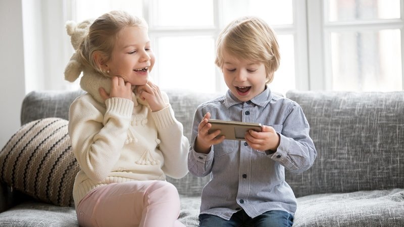 Excited kids using smartphone sitting together on sofa, happy little brother holding phone enjoying mobile app with smiling sister, cute children boy and girl having fun playing mobile game on gadget